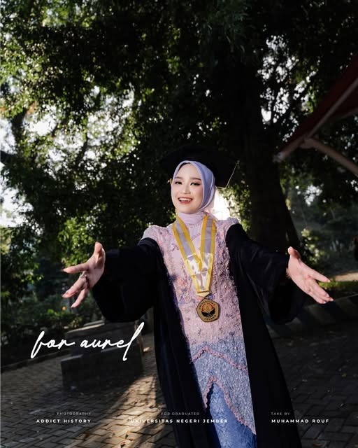 Wisudawati berhijab dengan toga dan medali, berpose di bawah pohon.